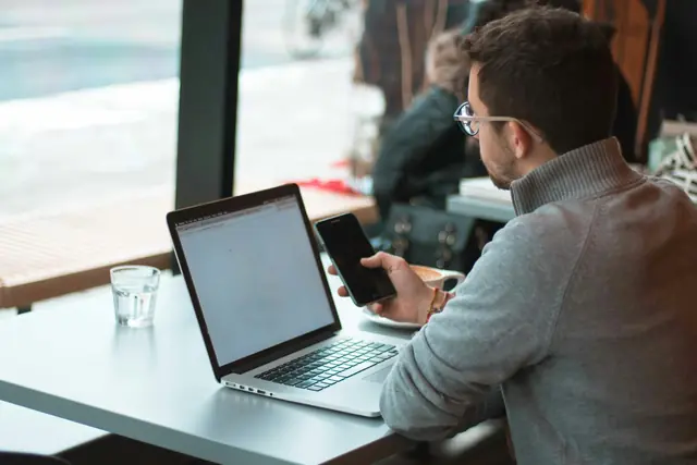 A man at a cafe table with a laptop, checking his phone